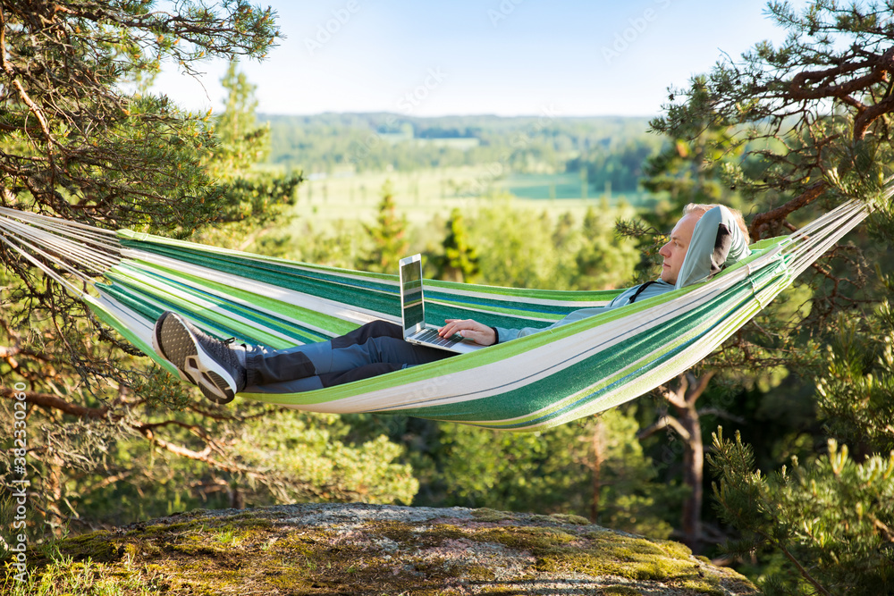 A man working on a laptop while lying in a hammock in the woods. Self-isolation, freelancing, remote work and distancing. Top view of Scandinavian landscape 