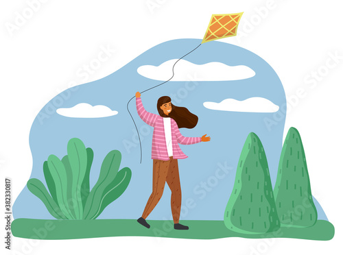 Smiling happy girl in park playing with kite flying in air in the wind. Child running holding thread in hand. Character with paper kite on background of green plants on grass and blue sky with clouds