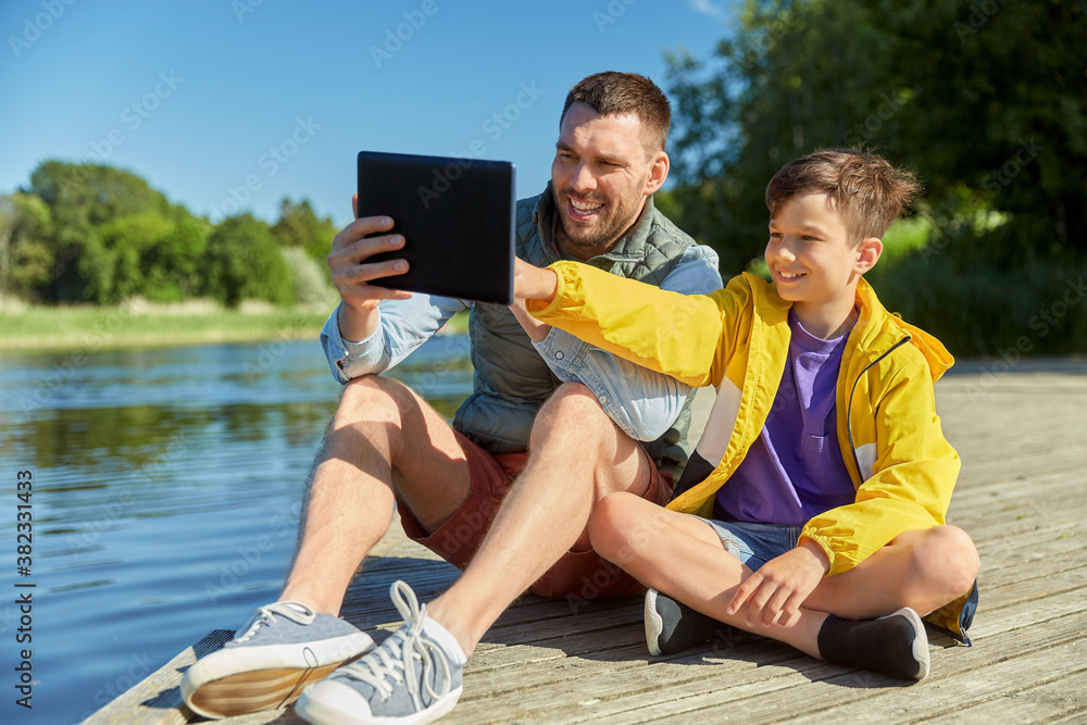 family, generation and technology concept - happy father and son with tablet pc computer sitting on river berth
