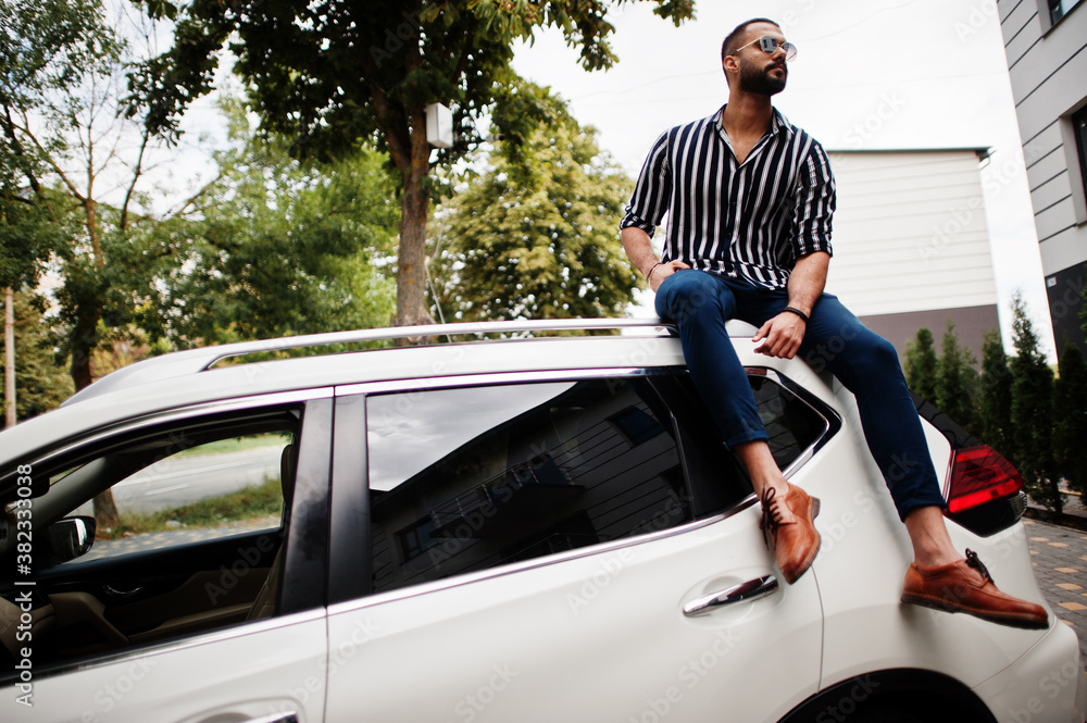 Successful arab man wear in striped shirt and sunglasses sitting on the roof of his white suv car.