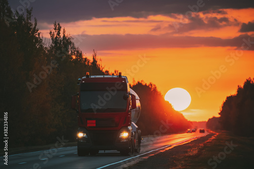 Red Truck Or Tractor Unit, Prime Mover, Traction Unit In Motion On Road, Freeway. Asphalt Motorway Highway Against Background Of Big Sunset Sun. Business Transportation And Trucking Industry.