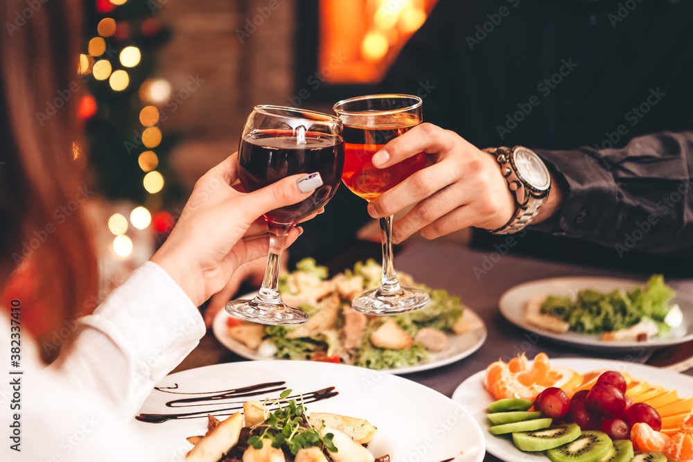 Close up of hands holding wine glasses and clinking against the backdrop of bright Christmas tree and burning fireplace, couple celebrating at Christmas table Close up of hands holding wine glasses and clinking against the backdrop of bright Christmas tree and burning fireplace, couple celebrating at Christmas table