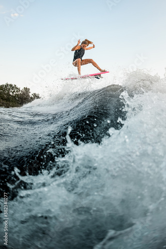 View on active young woman skillfully jumping over big splashing wave on surf style wakeboard.