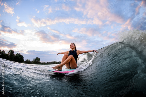 happy woman riding wave while sitting on surf style wakeboard with arms outstretched to the sides