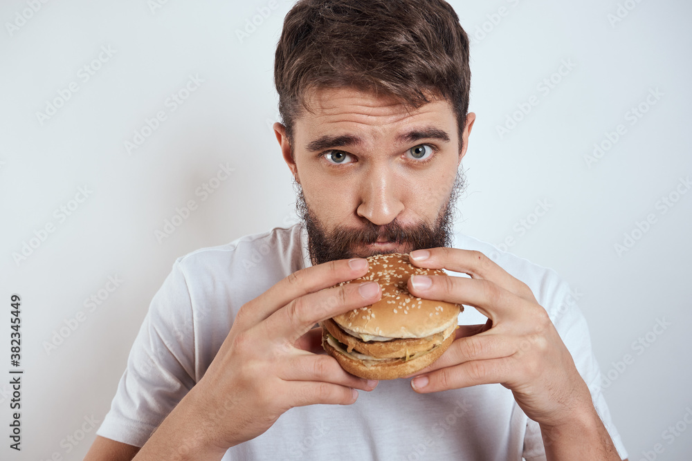 A man eating a hamburger on a light background in a white T-shirt cropped view close-up hunger fast food A man eating a hamburger on a light background in a white T-shirt cropped view close-up hunger fast food