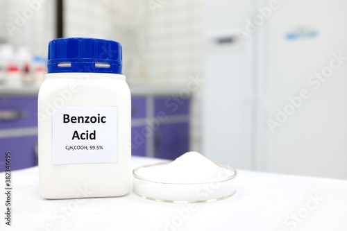 Selective focus of a bottle of pure benzoic acid chemical compound beside a petri dish with solid crystalline powder substance. White Chemistry laboratory background with copy space. photo