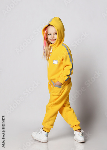 Smiling blonde kid girl with braids in stylish yellow jumpsuit with her hood on stands back to camera and looks at us