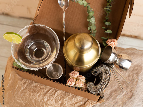Bar cocktail party, bartender stuff accessories delivery parcel in a natural brown craft cardboard box. A gift to mixology lover. Beige brick wall background. Flat lay top view. Retro hipster colors