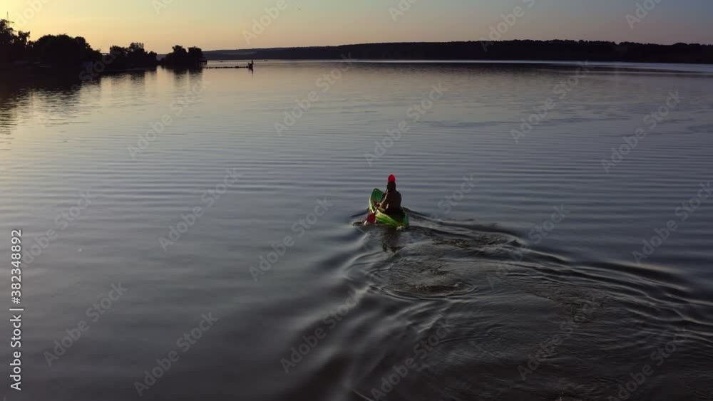 Young man kayaks on lake at sunset. Alone man riding on a rowing boat on the lake at dusk time. Man in a boat with oars in the evening. Aerial view. Young man kayaks on lake at sunset. Alone man riding on a rowing boat on the lake at dusk time. Man in a boat with oars in the evening. Aerial view.