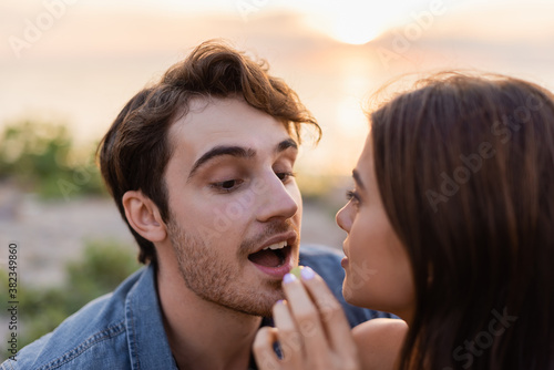 Selective focus of young woman feeding boyfriend with grape on beach at sunset