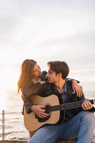 Young woman in leather jacket embracing boyfriend playing acoustic guitar near sea at sunset © LIGHTFIELD STUDIOS