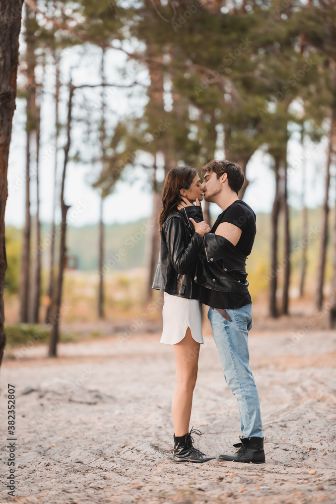 Side view of man in leather jacket kissing brunette girlfriend on sand in forest Side view of man in leather jacket kissing brunette girlfriend on sand in forest