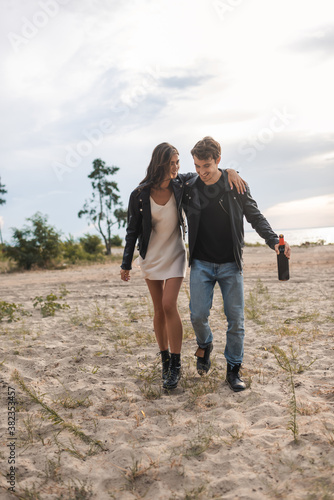 Couple in leather jackets embracing while walking with bottle of wine on sandy beach