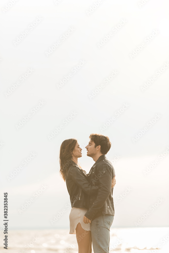 Side view of woman in leather jacket hugging boyfriend with sea and sky at background