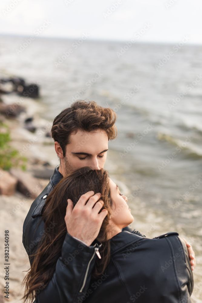 Young man touching hair of brunette girlfriend in leather jacket near sea
