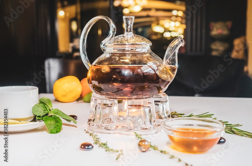 Aromatic hot green tea in a glass teapot with a warm blurry background with honey and fresh mint leaves. Served table with tea in a cafe. Kettle warmer