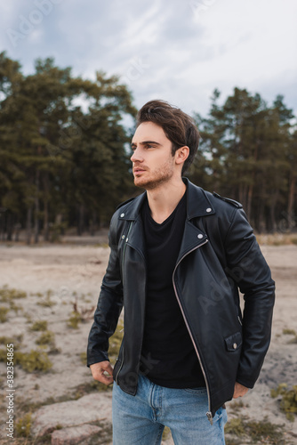 Young man in leather jacket looking away near forest at background