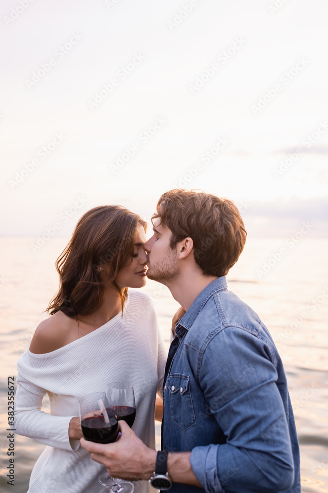 Selective focus of young man kissing nose of girlfriend with glass of wine near sea