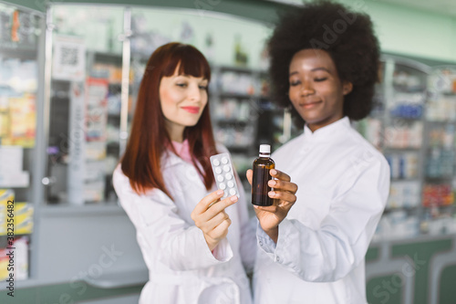 Two multiethnic pharmacists women, African and Caucasian, working in drugstore, checking medicines quality at hospital pharmacy, showing to camera syrup bottle and pill blister. Focus on drugs
