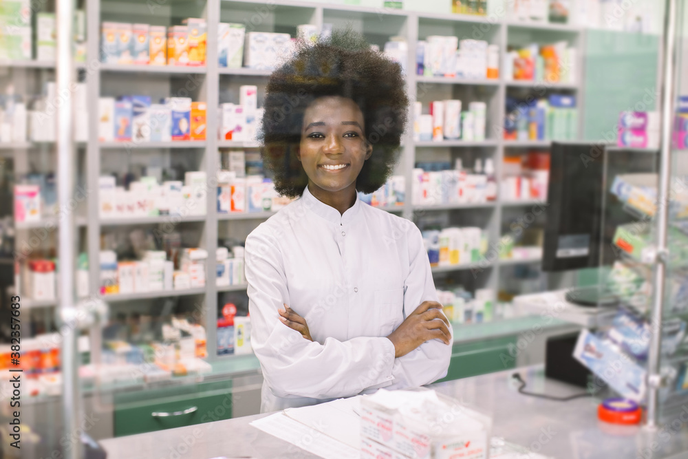 Happy young dark skinned woman pharmacist, posing to camera with smile, standing with arms crossed over drugstore background. Medicine, pharmacy, people, health care and pharmacology concept