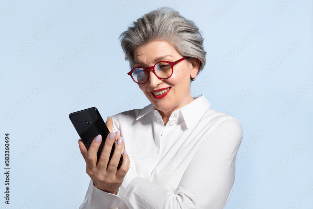 Friendly smiling senior woman in glasses with smartphone using mobile apps on blue background. Positive elderly female with gray hair in white shirt shopping online. Easy technology concept Friendly smiling senior woman in glasses with smartphone using mobile apps on blue background. Positive elderly female with gray hair in white shirt shopping online. Easy technology concept
