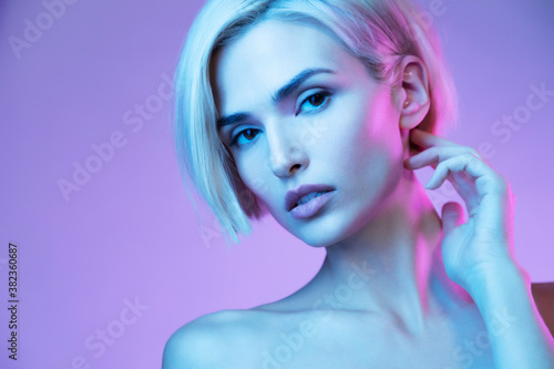 Young blond female with stylish fashionable haircut and naked shoulders in neon colorful studio light on lilac background. Close up beauty portrait of girl with natural make up and perfect skin.