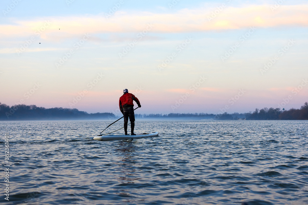 Back view on man rowing on SUP (stand up paddle board) in autumn Danube river at foggy morning against the backdrop of autumn trees without leaves Back view on man rowing on SUP (stand up paddle board) in autumn Danube river at foggy morning against the backdrop of autumn trees without leaves