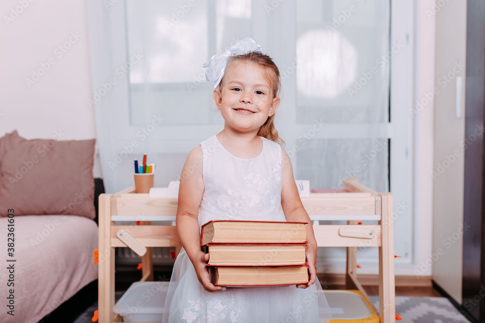 Little student holding big red books near school desk. Back to school. Little girl studies at home. Little student holding big red books near school desk. Back to school. Little girl studies at home.