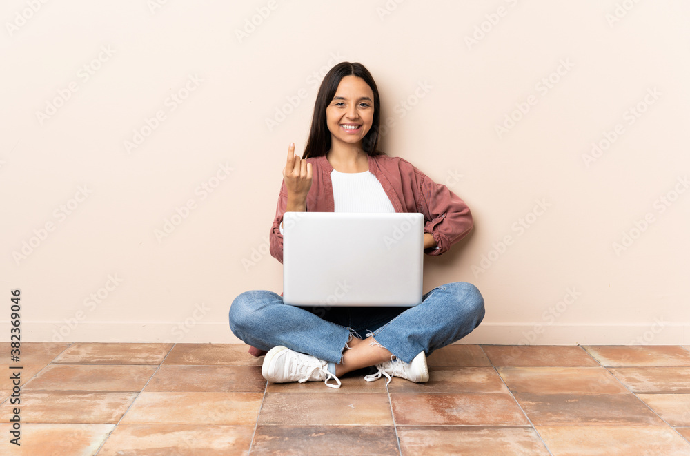 Young mixed race woman with a laptop sitting on the floor doing coming gesture Young mixed race woman with a laptop sitting on the floor doing coming gesture