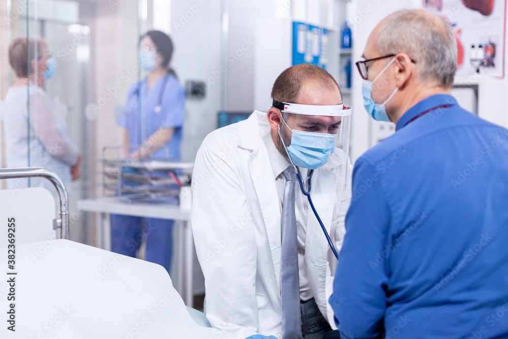 Fototapeta premium Medical practitioner checking heart beat of senior patient during consultation with stethoscope and wearing face mask againts covid-19. Medical examination for infections, disease and diagnosis.