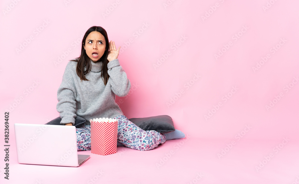Young mixed race woman eating popcorn while watching a movie on the laptop listening to something by putting hand on the ear Young mixed race woman eating popcorn while watching a movie on the laptop listening to something by putting hand on the ear