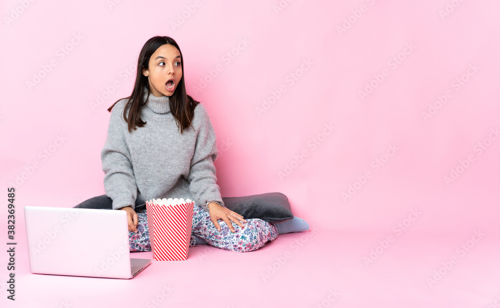 Young mixed race woman eating popcorn while watching a movie on the laptop doing surprise gesture while looking to the side Young mixed race woman eating popcorn while watching a movie on the laptop doing surprise gesture while looking to the side
