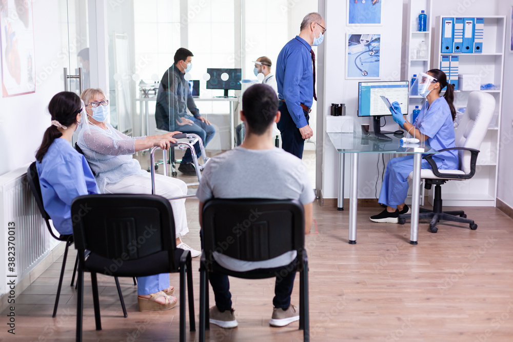Nurse taking notes on clipboard wearing visor and sterile gloves against coronavirus while questioning elderly man in hospital waiting area. Social distancing, medic, covid-19, man, worried, Nurse taking notes on clipboard wearing visor and sterile gloves against coronavirus while questioning elderly man in hospital waiting area. Social distancing, medic, covid-19, man, worried,