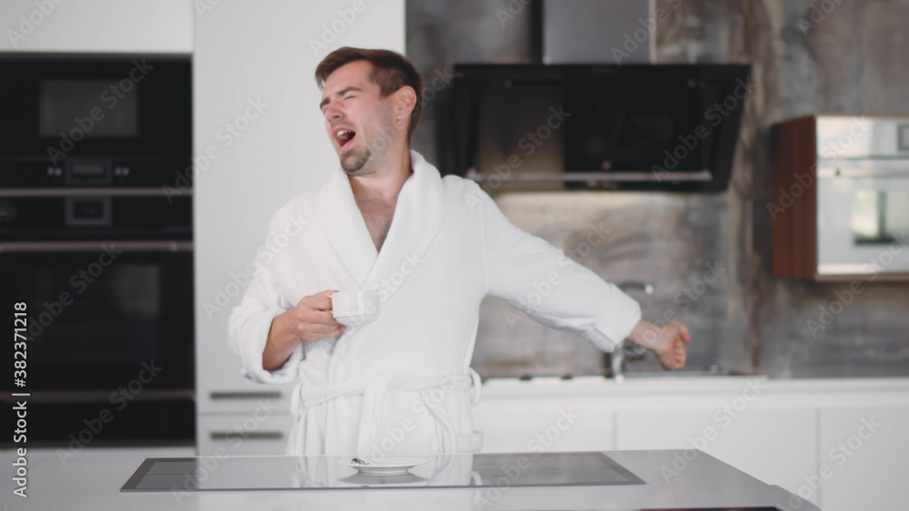 Handsome man in bathrobe yawning and stretching at kitchen enjoying cup of coffee in morning. Portrait
