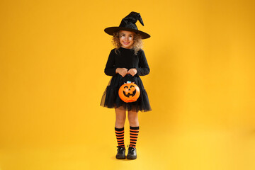 Cute little girl with pumpkin candy bucket wearing Halloween costume on yellow background