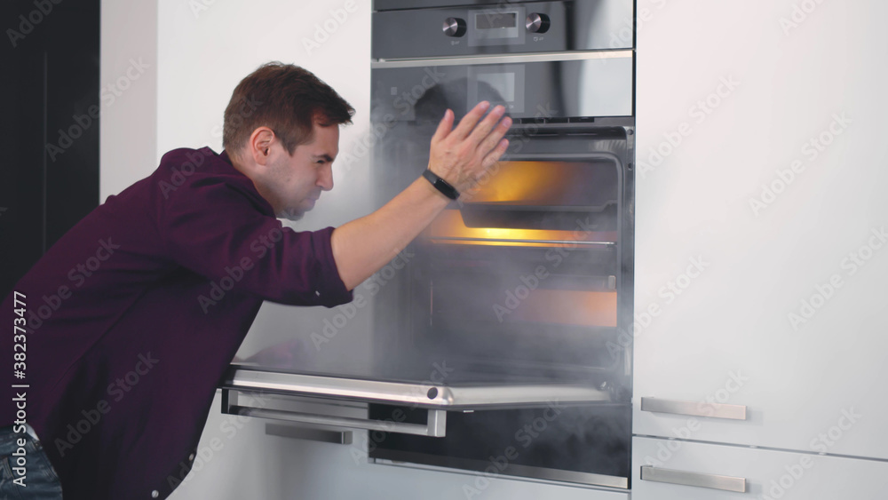 Young Shocked Man Opening Oven Filled With Smoke In Kitchen Young Shocked Man Opening Oven Filled With Smoke In Kitchen