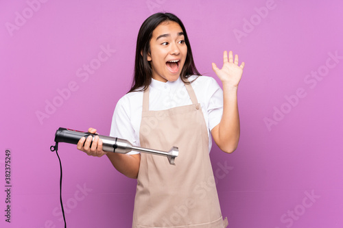 Indian woman using hand blender isolated on purple background shouting with mouth wide open