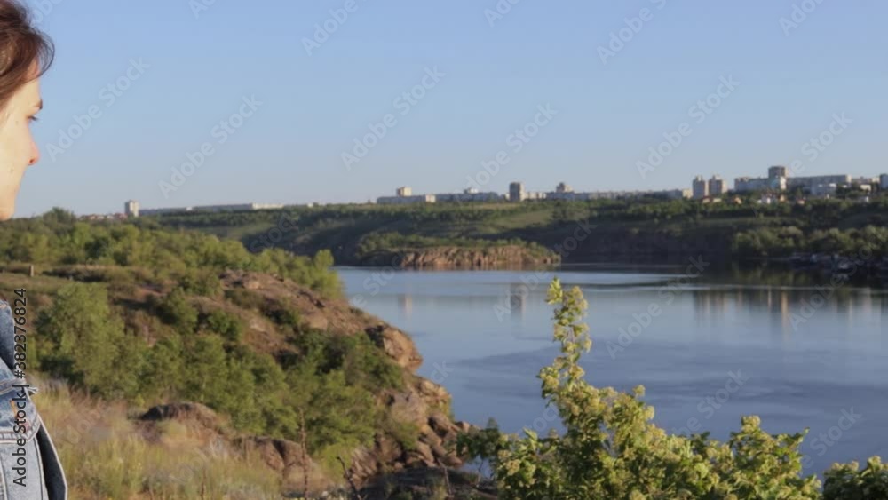A girl in a denim jacket with a backpack admires the beautiful view of the river and the city in the distance. A girl in a denim jacket with a backpack admires the beautiful view of the river and the city in the distance.