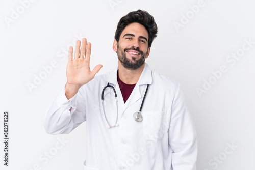 Young handsome man with beard over isolated white background wearing a doctor gown and saluting