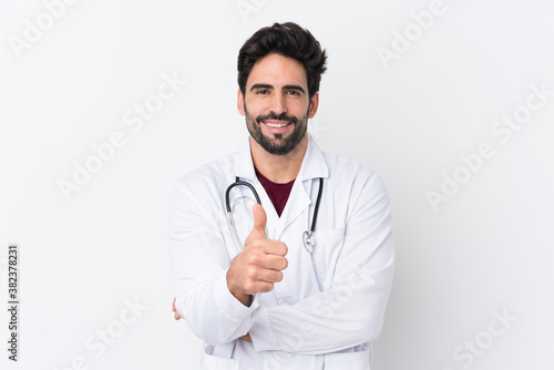 Young handsome man with beard over isolated white background wearing a doctor gown and with thumb up