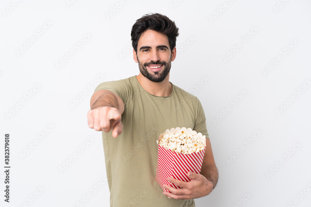 Young handsome man with beard over isolated white background holding a big bucket of popcorns while pointing front