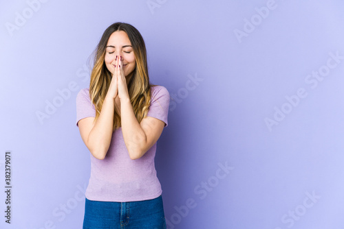 Young caucasian woman isolated on purple background holding hands in pray near mouth, feels confident. © Asier