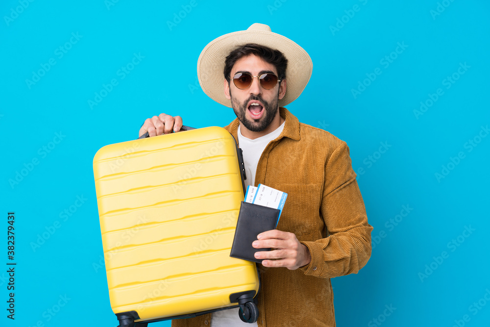 Young handsome man with beard over isolated blue background in vacation with suitcase and passport Young handsome man with beard over isolated blue background in vacation with suitcase and passport