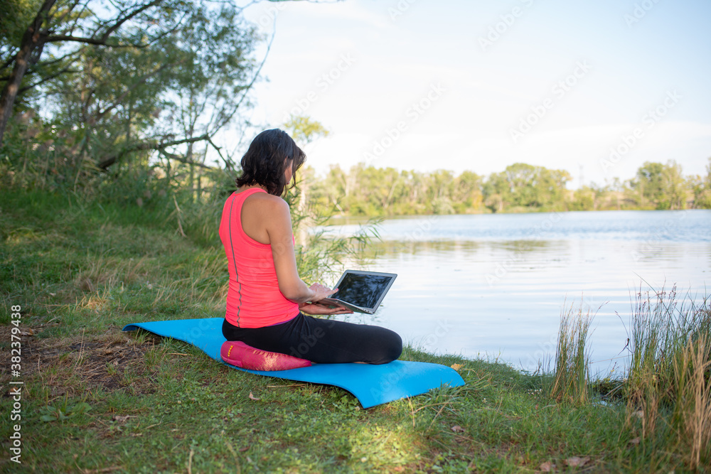 young woman practicing yoga and meditation on a yoga mat outdoor with a laptop