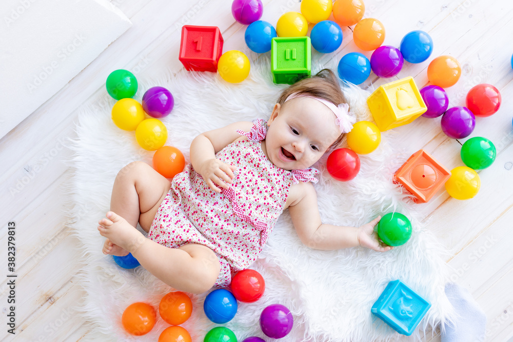 Obraz premium a beautiful little baby girl is lying in red clothes on a white Mat among toys balls and cubes