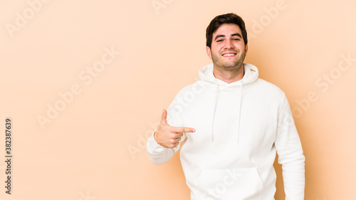 Young man isolated on beige background person pointing by hand to a shirt copy space, proud and confident © Asier