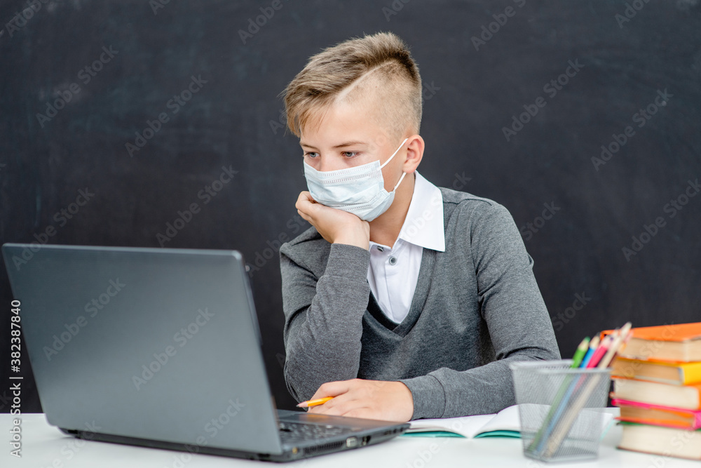 Teen boy wearing mask uses laptop at school near blackboard during corona virus and flu outbreak