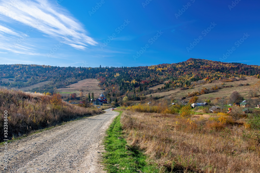 Naklejka premium Dirt road to church in small village among colorful hills in Carpathian Mountains, Ukraine at sunny autumn day