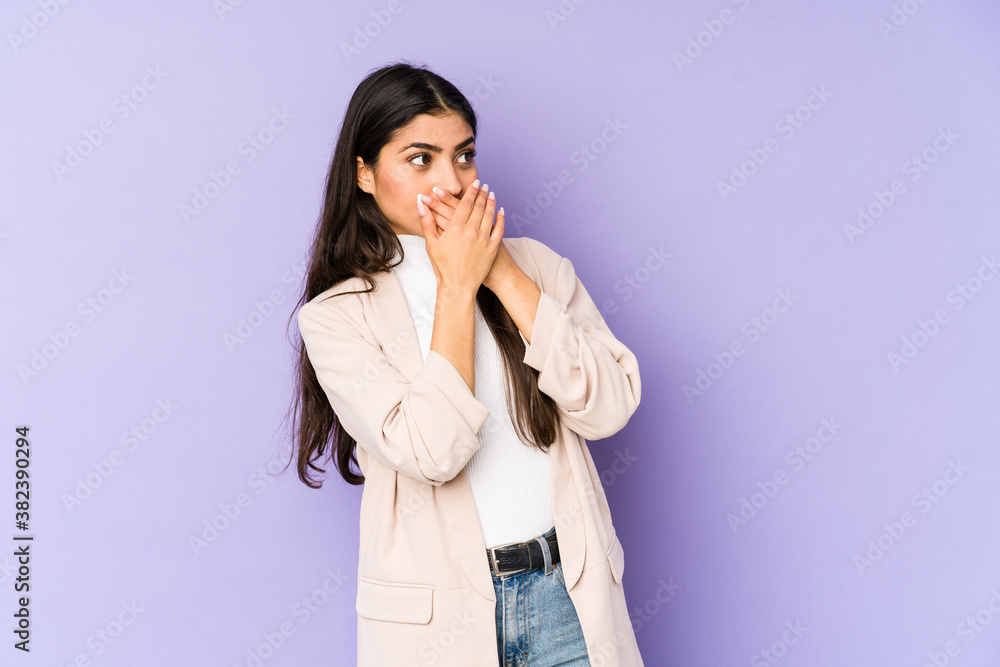 Young indian woman isolated on purple background thoughtful looking to a copy space covering mouth with hand. Young indian woman isolated on purple background thoughtful looking to a copy space covering mouth with hand.