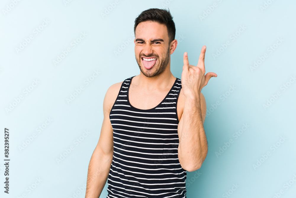 Young caucasian man isolated on blue background showing rock gesture with fingers Young caucasian man isolated on blue background showing rock gesture with fingers